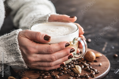 Women's hands in a warm sweater are holding a cup of fragrant latte. The nails are coated with brown nail polish. Coffee manicure. Breakfast or a break in a cozy cafe