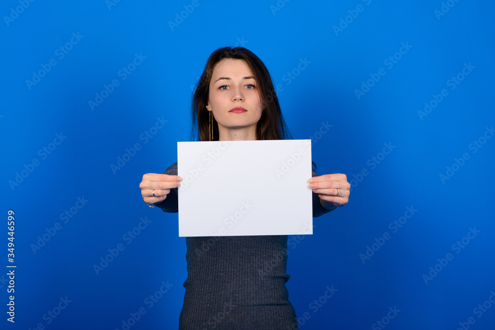 Young woman in grey shirt holding a sheet of paper and looking at the camera, smiling. Blue background isolated Young woman in grey shirt holding a sheet of paper and looking at the camera, smiling. Blue background isolated