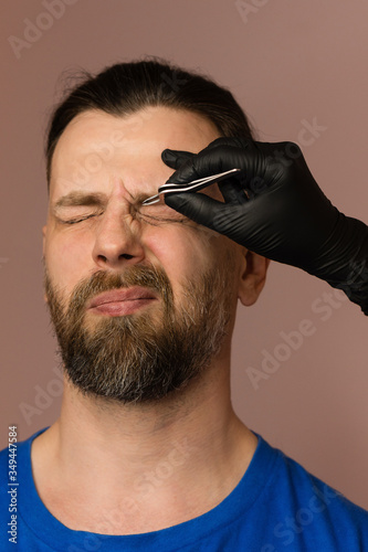 Professional barber doing threading procedure and correcting shape of eyebrows to young male client with tweezer in barber shop. Barber at work. Plucking brows. Customer hurt