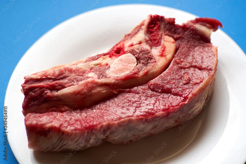 Raw veal steak on a white plate on a blue background.