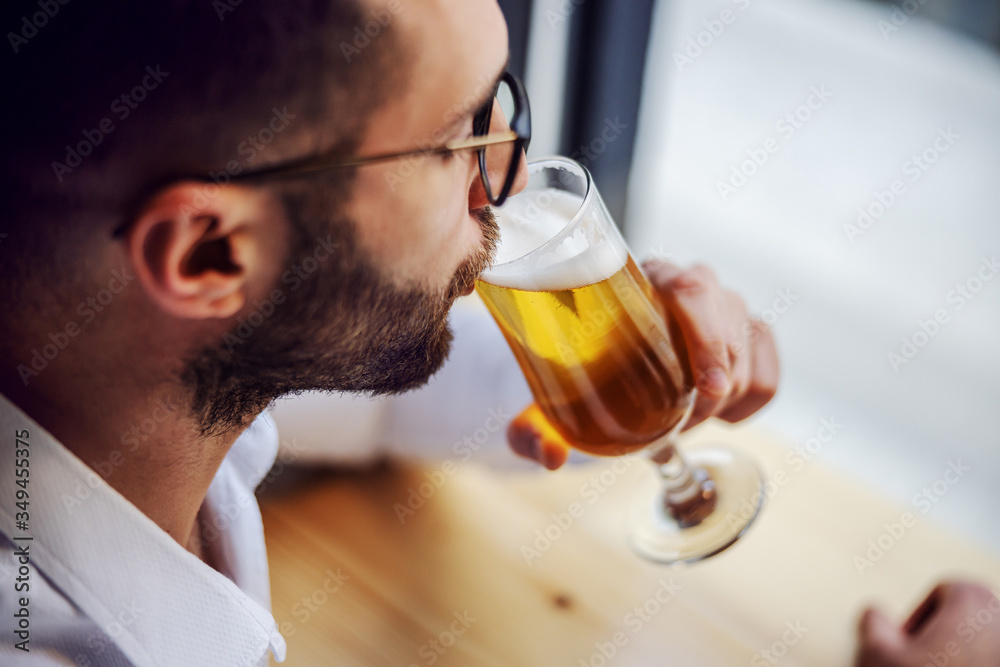 Closeup of handsome unshaven man in shirt sitting in pub next to window, drinking beer and relaxing after work. Closeup of handsome unshaven man in shirt sitting in pub next to window, drinking beer and relaxing after work.