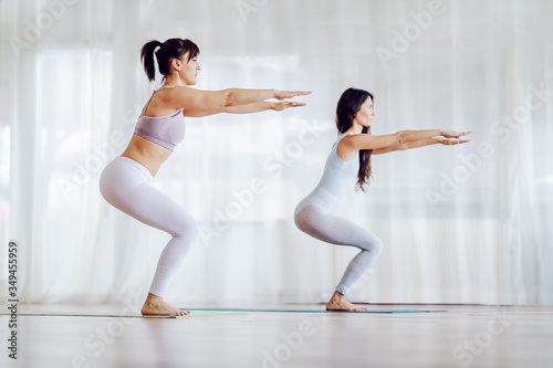 Side view of two focused girls in shape in Awkward yoga pose. Yoga studio interior.