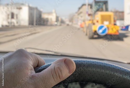 view of the driver hand on the steering wheel of a car passing through the courtyard of the street where the tractor stands  road signs and roadway repairs and maintenance are being carried out