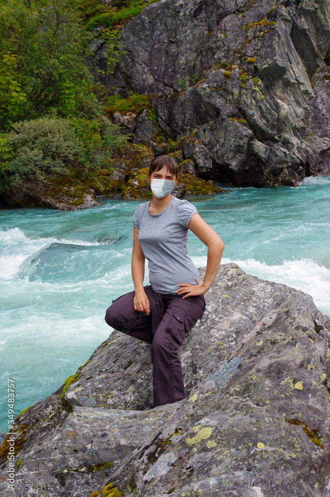 Woman in a medical mask and a waterfall in Norway. Travel and tourist vacation concept during Coronavirus Covid-19.