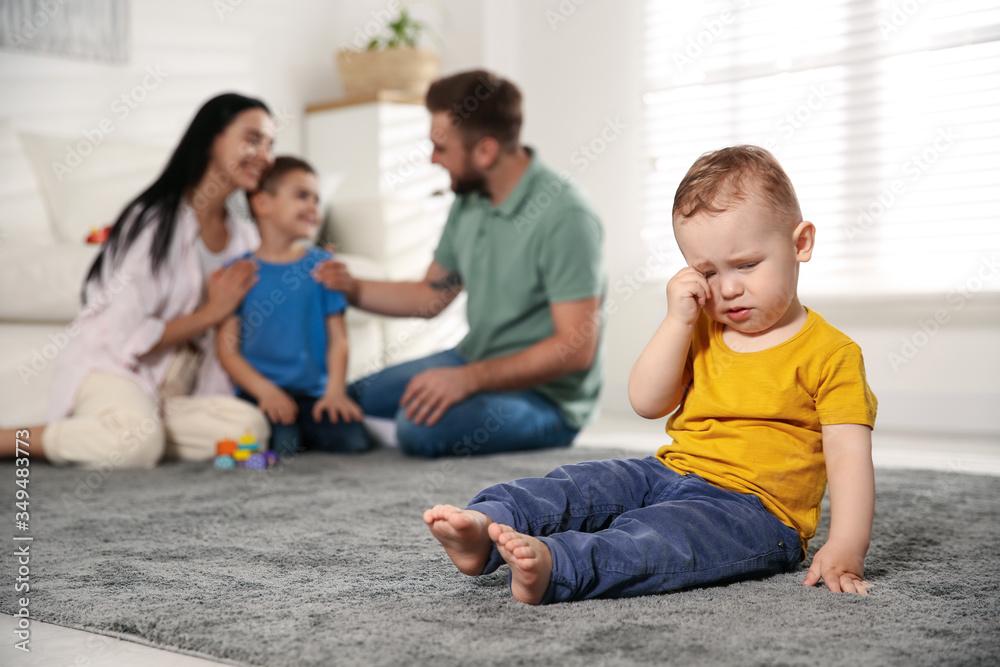 Unhappy baby sitting alone on floor while parents spending time with his elder brother at home. Jealousy in family Unhappy baby sitting alone on floor while parents spending time with his elder brother at home. Jealousy in family