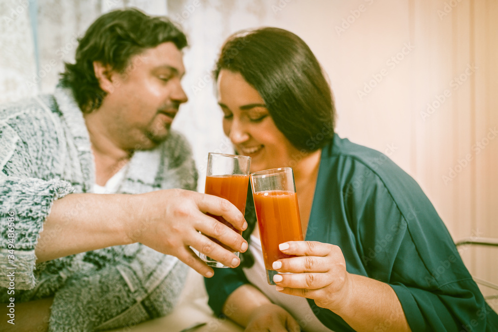 Smiling overweight couple combined glasses with orange juice. Man says a festive toast while sitting at the kitchen table Smiling overweight couple combined glasses with orange juice. Man says a festive toast while sitting at the kitchen table