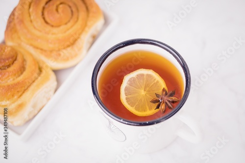 A cup of lemon tea with anise star and homemade cinnabons, cinnamon rolls buns on white marble background