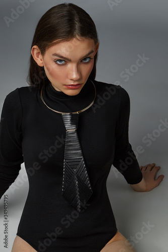 A ravishing brunette lady in a black bodysuit and a trendy choker is sitting on the gray floor. The carcanet is made as golden wires with hanging black tie consists of weaving leather straps.  photo