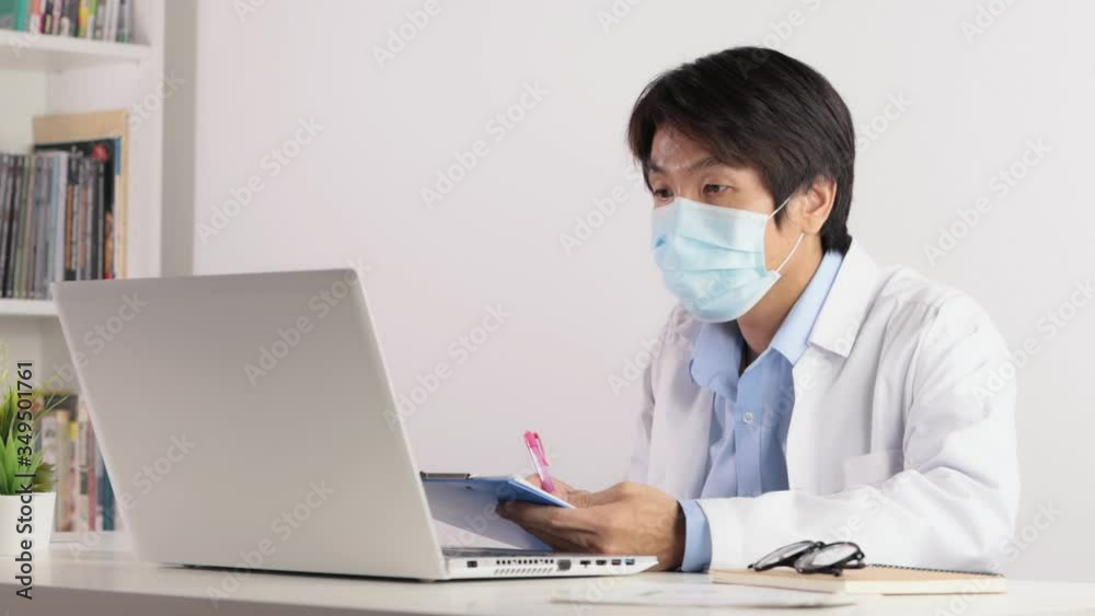 Asian Male Doctor Wear Mask and White Coat or Gown Writing Report and Video Call with Patient. Asian doctor see laptop monitor in serious emotion Asian Male Doctor Wear Mask and White Coat or Gown Writing Report and Video Call with Patient. Asian doctor see laptop monitor in serious emotion