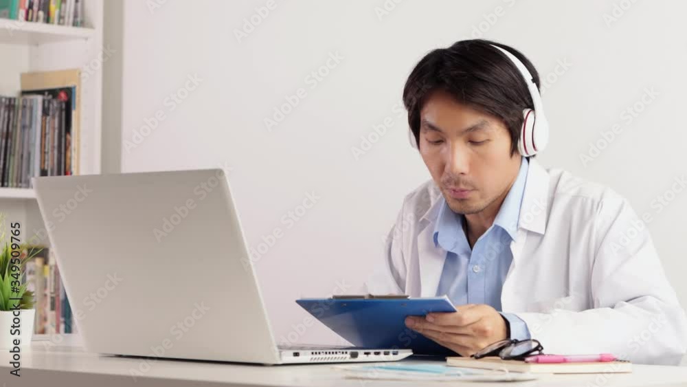 Asian Male Doctor Wear Headphone See Report Working With Laptop Pass Video Call or Video Conference System. Asian male doctor in serious emotion Asian Male Doctor Wear Headphone See Report Working With Laptop Pass Video Call or Video Conference System. Asian male doctor in serious emotion