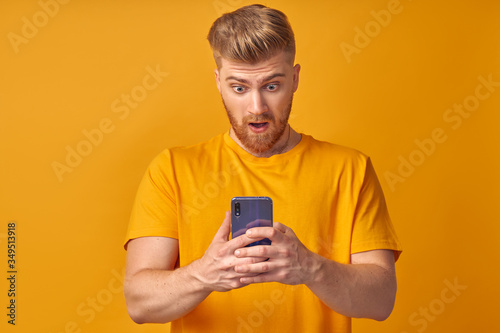 Portrait of handsome man looks shocked in smart phone, sees calendar date, finds out that he can`t manage to go on meeting, types message of refusal, yellow background. People, emotions and technology © antianti