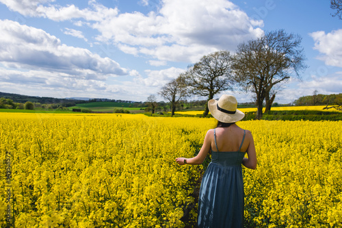 Girl from behind wearing a blue dress and a hat contemplating a beautiful yellow rapeseed flowers field in a sunny day