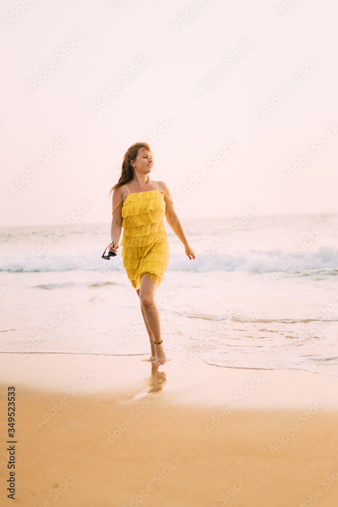 Goa, India. Young Caucasian Woman In Yellow Dress Walking On Seashore And Enjoying Life In Summer Sunlight