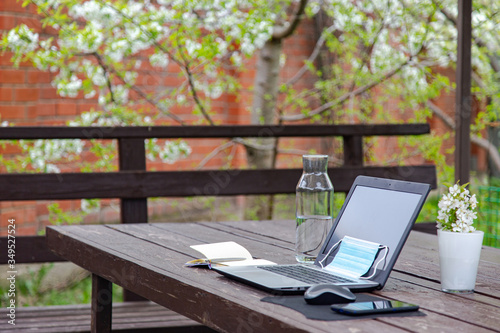 remote work or distant study from home during the coronavirus pandemic. Medical mask, laptop, mouse, notepad, mobile phone on a brown wooden table in garden. Blooming tree.