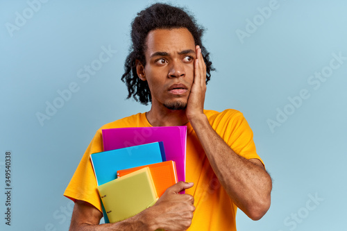 Excited shocked young African-American student holding notebooks and worrying about exams on blue background. Concept of online courses and preparation for entering university. Advertising space