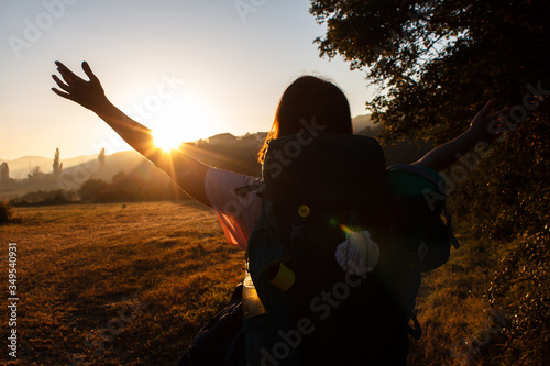 Girl raising arms up and meeting beautiful sunrise on the hills in Spain. Girl hiker meets the dawn. Camping backpack with a shell, which is a symbol of the Way of Saint James. Walking camino. photo