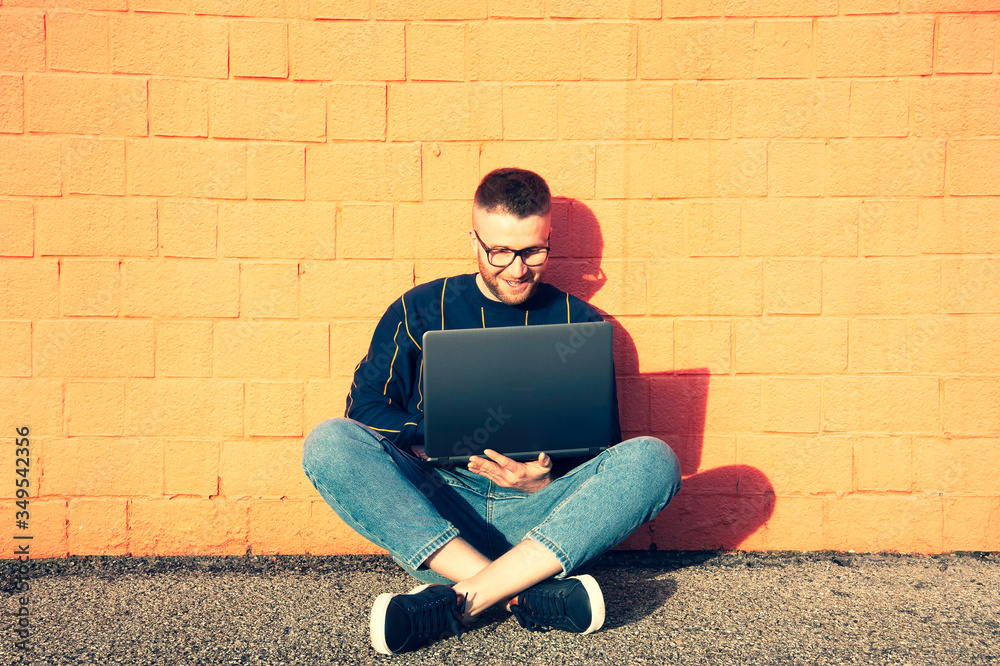 Young caucasian man in casual-wear sitting cross-legged on ground and working on laptop - Freelancer working outside typing on laptop computer, Smart working concept - Red wall on the background