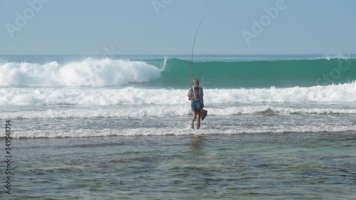 local fisherman holds long rod in hand standing in waving blue ocean against clear sky on hot tropical day slow motion. Concept tropical seascape travel photo