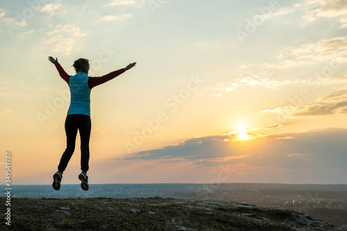 Silhouette of a woman hiker jumping alone on empty field at sunset in mountains. Female tourist raising her hands up in evening nature. Tourism, traveling and healthy lifestyle concept.