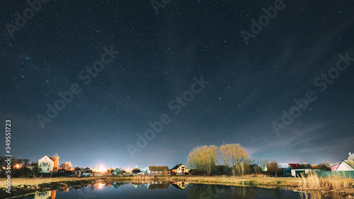 Belarus, Eastern Europe. Night Sky Stars Above Countryside Landscape With Lake Coast And Small Town Or Village. Natural Starry Sky Above Pond And Houses In Early Spring Night. Panorama Panoramic View.