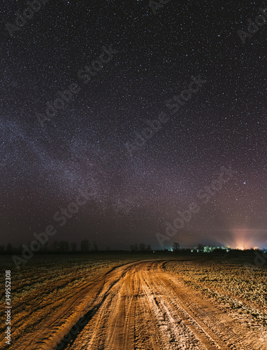 Night Starry Sky With Glowing Stars Above Countryside Road Landscape. Milky Way Galaxy And Rural Field Meadow