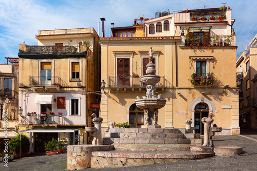 Fountain and typical houses with restaurants on the square Piazza Duomo in Taormina on sunny morning, Sicily, Italy