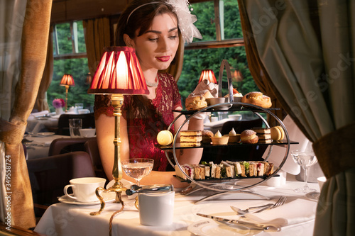 Vintage  beautiful woman in red dress enjoying afternoon tea in train carriage with cakes  sandwiches and tea
