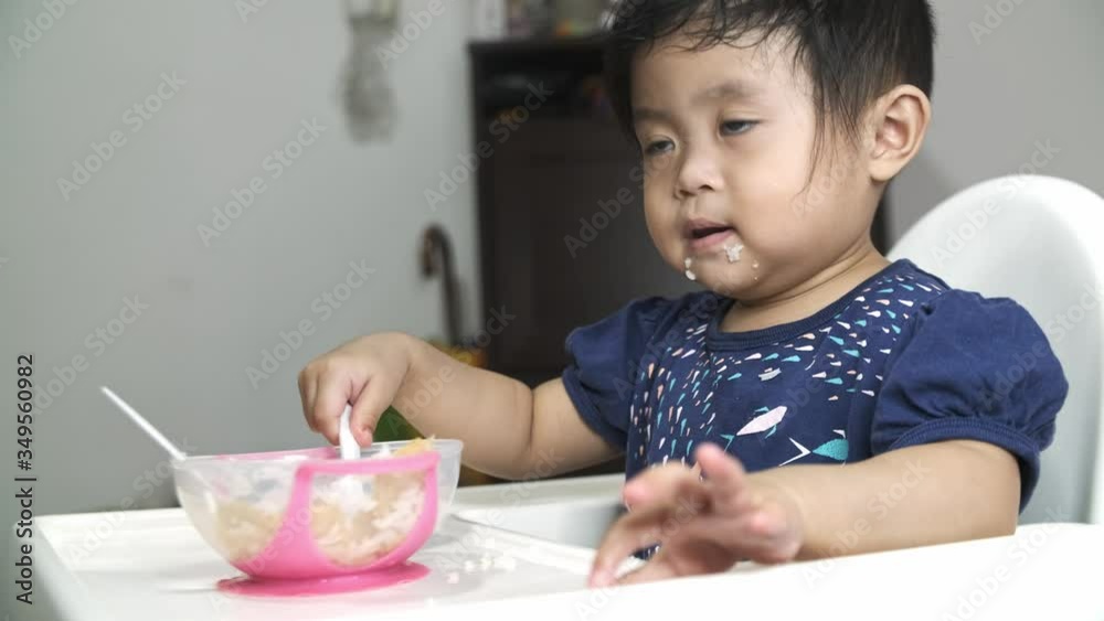 Cute Malaysian baby girl sitting at baby high chair eating lunch, medium shot Cute Malaysian baby girl sitting at baby high chair eating lunch, medium shot