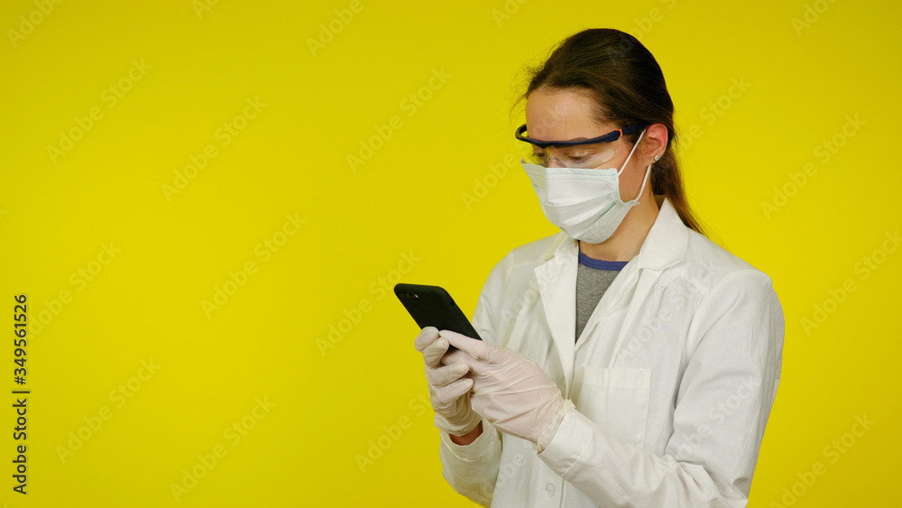 Young doctor in a protective medical mask, glasses and latex gloves looks at the smartphone. Nurse in a white coat on yellow background is looking for information in a smartphone