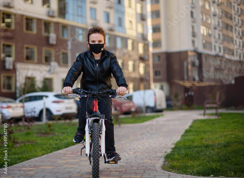 Boy in a leather jacket wearing a black hygyenic medical mask outdoors in the yard, riding a bike. High-rise building on the background