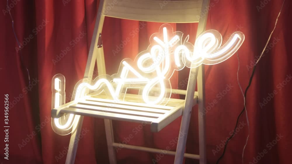 title that says imagine made of yellow neon light on wooden chair. festive decoration of interior. red curtain on background. illuminated letters of elecrical tubes shine bright in the evening