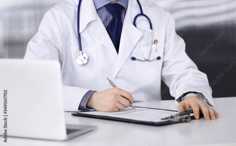 Unknown male doctor sitting and working with clipboard in clinic at his working place, close-up. Young physician at work. Perfect medical service, medicine concept Unknown male doctor sitting and working with clipboard in clinic at his working place, close-up. Young physician at work. Perfect medical service, medicine concept
