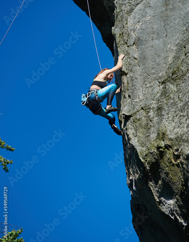 Lady mountaineer in sportswear climbing rock under blue sky. Young woman climber ascending high mountain and trying to reach mountaintop. Concept of mountaineering, alpine climbing and extreme sports.