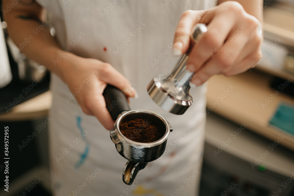A close-up photo of female hands holding a metal tamper and a portafilter with coffee in a coffee shop. A barista preparing for pressing ground coffee for brewing espresso or americano in a cafe. A close-up photo of female hands holding a metal tamper and a portafilter with coffee in a coffee shop. A barista preparing for pressing ground coffee for brewing espresso or americano in a cafe.