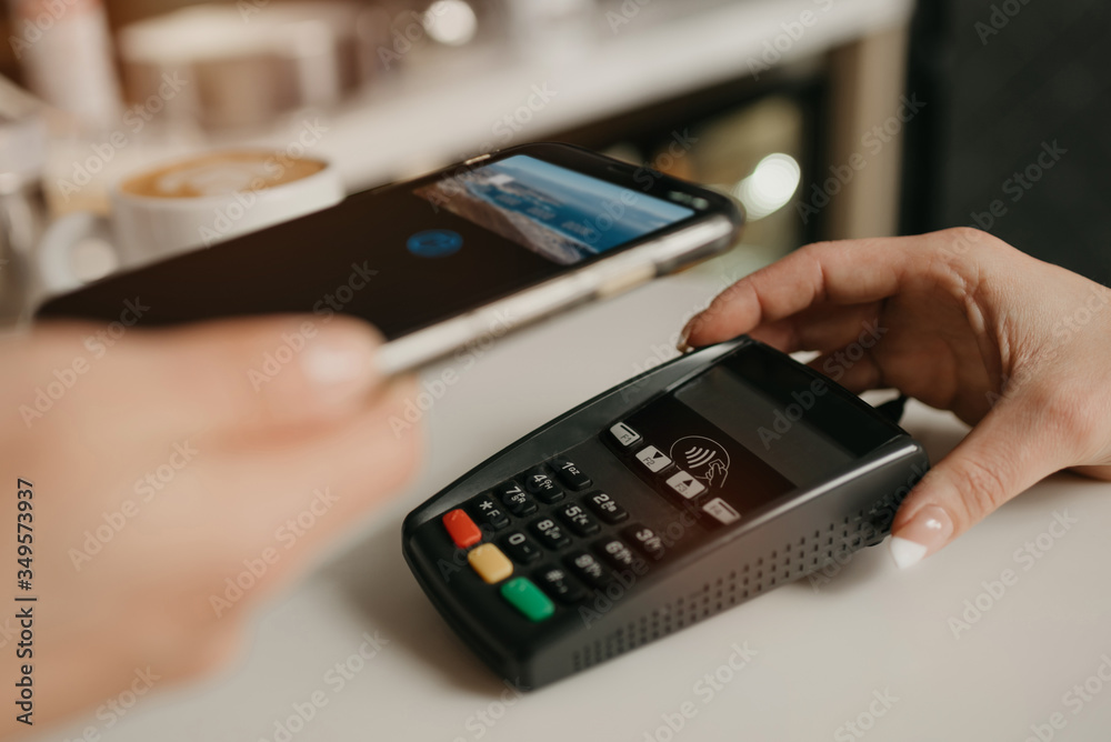 A lady paying for her latte with a smartphone by contactless NFC technology in a cafe. A female barista holds out a terminal for paying to a client in a coffee shop. A lady paying for her latte with a smartphone by contactless NFC technology in a cafe. A female barista holds out a terminal for paying to a client in a coffee shop.