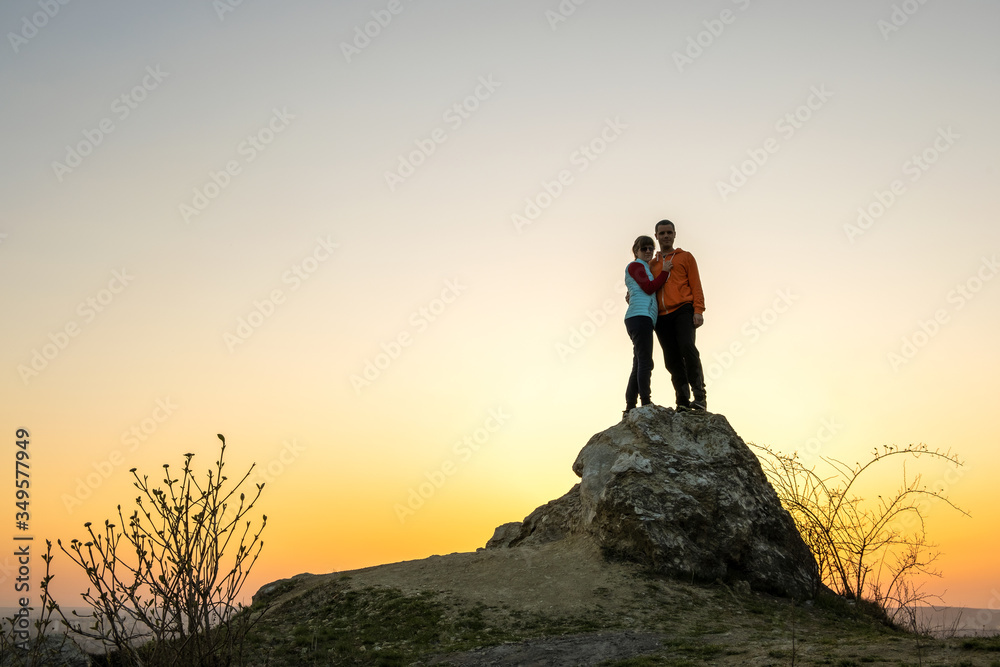 Man and woman hikers standing on a big stone at sunset in mountains. Couple together on a high rock in evening nature. Tourism, traveling and healthy lifestyle concept.