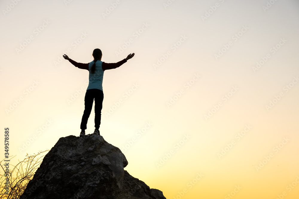 Silhouette of a woman hiker standing alone on big stone at sunset in mountains. Female tourist raising her hands up on high rock in evening nature. Tourism, traveling and healthy lifestyle concept. Silhouette of a woman hiker standing alone on big stone at sunset in mountains. Female tourist raising her hands up on high rock in evening nature. Tourism, traveling and healthy lifestyle concept.