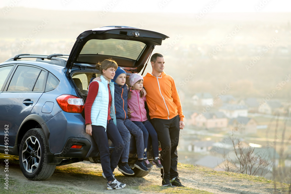Happy family standing together near a car with open trunk enjoying view of rural landscape nature. Parents and their kids leaning on vehicle luggage compartment. Weekend travel and holidays concept.