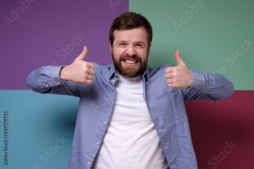 Gloating Caucasian man makes an insincere approving gesture with hands, raises thumbs up and grins teeth, squinting eyes.