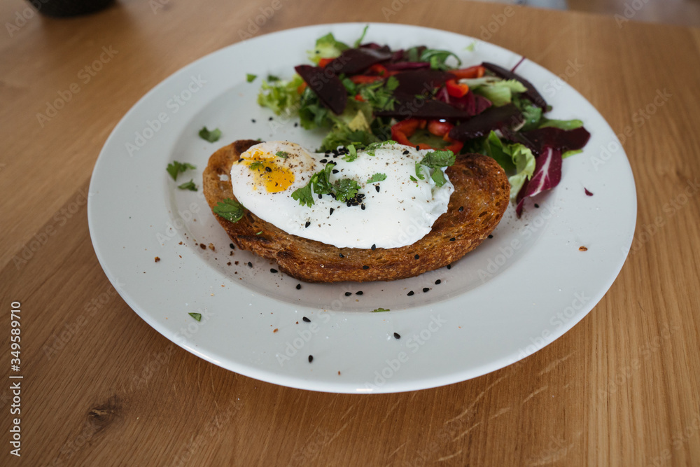 Poached egg on a piece of bread with green salad on a plate and on the wooden table. Healthy Breakfast