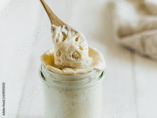 Ready for making bread wheat sourdough starter dripping from wooden spoon. Glass jar with sourdough starter 100 percent hydration on white wooden background. Copy space. Homemade breadmaking concept