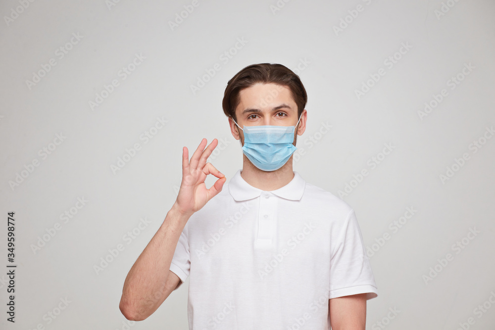 A man in a white T-shirt of European appearance in a medical mask, shows a thumb up. In the mask you are protected from covid 19 viruses.