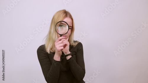 Young Blonde Woman In Black Sweater With Stylish Watch On White Background, Focused Girl Looks Through A Magnifying Glass, Big Eye. The Concept Of Research, Study, And Smart People