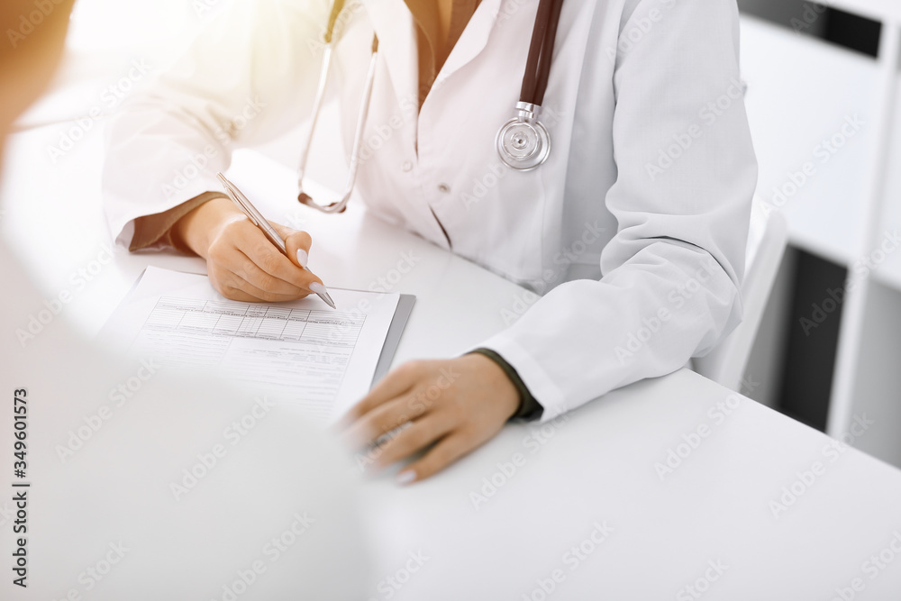 Unknown woman-doctor and female patient sitting and talking at medical examination in sunny clinic, close-up. Therapist filling up medication history record. Medicine concept Unknown woman-doctor and female patient sitting and talking at medical examination in sunny clinic, close-up. Therapist filling up medication history record. Medicine concept