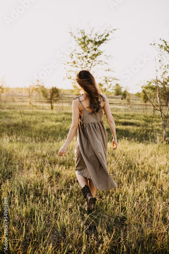 Young brunette woman with long wavy hair, wearing vintage sundress, walking in a golden field at the sunset.