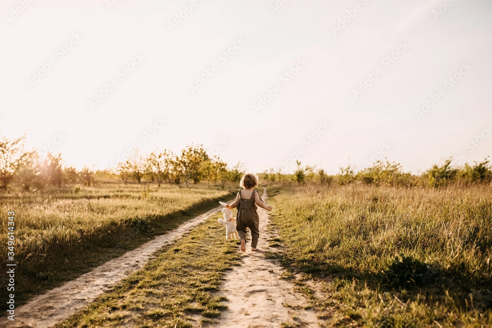 Little blonde boy with curly hair, wearing vintage jumpsuit, running on a dirt path, barefoot, at sunset, holding a plush rabbit toy.