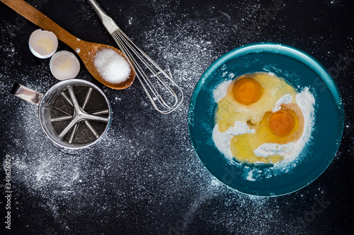 flour and eggs in blue case with beater, sieve, egg shells and sugar in wooden spoon. recipe of cake. cake ingredients