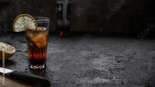 Bild auf Leinwand A glass of rum with cola full of ice with a slice of lemon on a dark gray bar table in grunge style