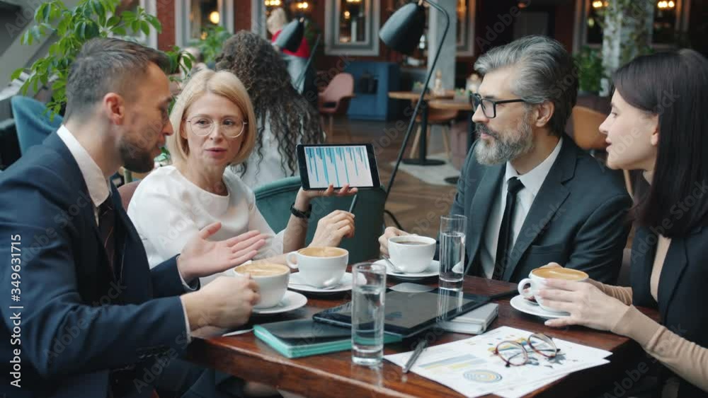 Mature business lady is using tablet during negotiations with business partners in restaurant talking showing data on gadget screen. People and devices concept. Mature business lady is using tablet during negotiations with business partners in restaurant talking showing data on gadget screen. People and devices concept.