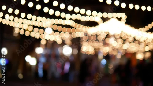 Glittering blurred bokeh of festival decorative string lights hang and glow outdoors at night with blurrry crowd people moving in festive Christmas market festival, abstract background.